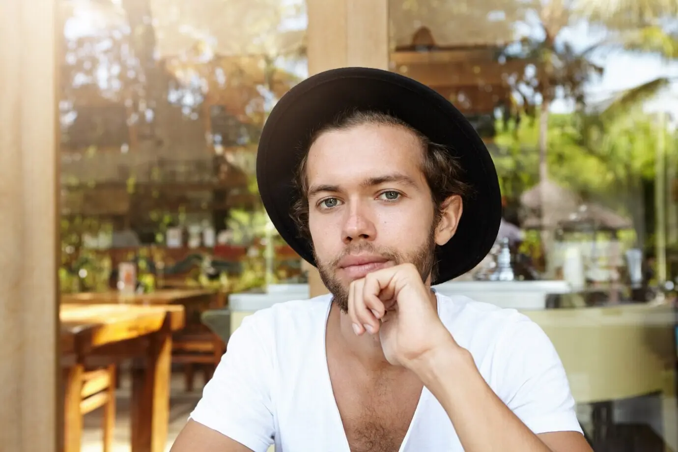 Porträtaufnahme eines modischen, attraktiven, jungen Studenten mit stylischem Bart, der einen schwarzen Hut und ein Shirt mit V-Ausschnitt trägt und lächelnd schaut.