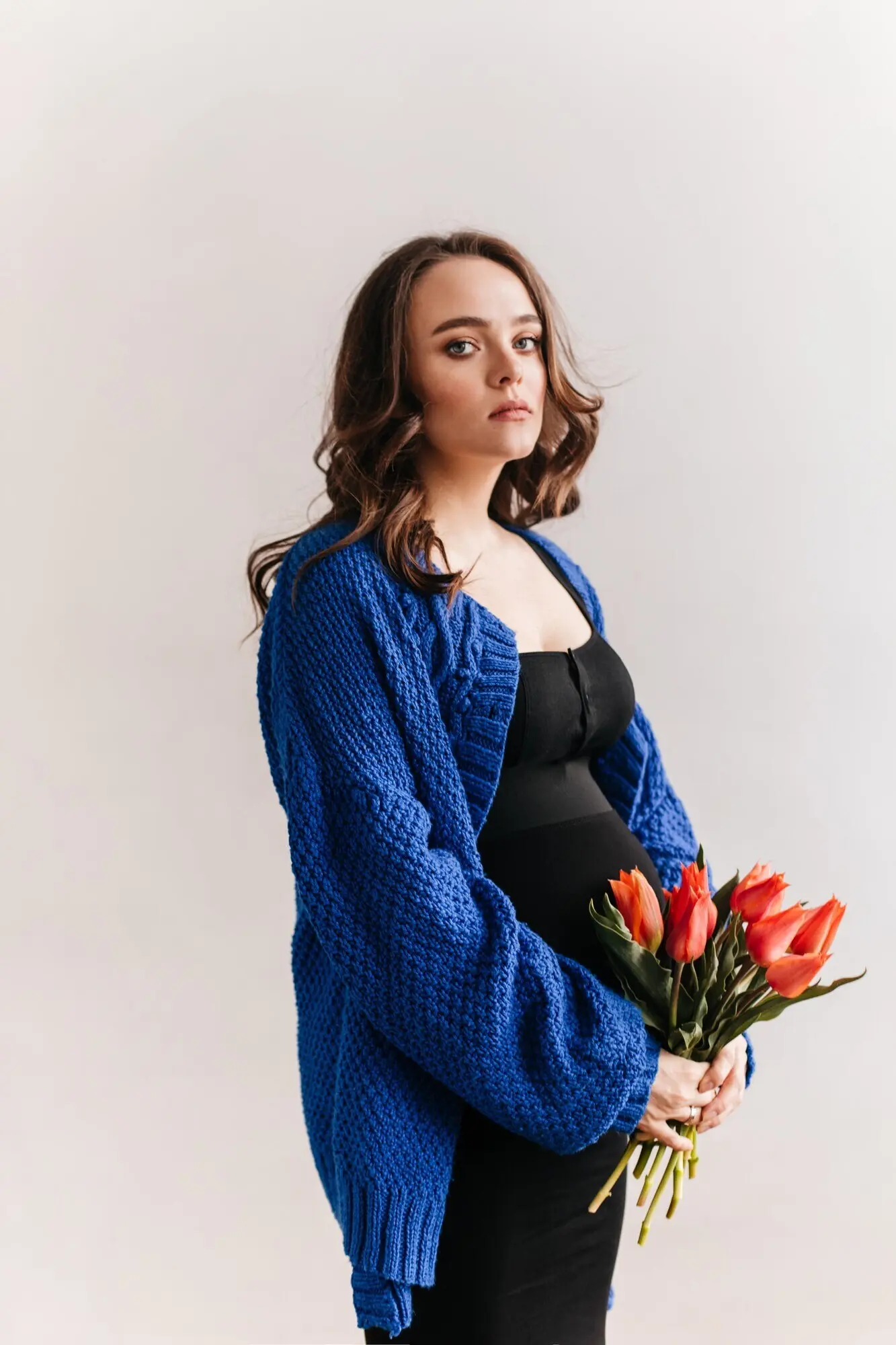Lockige brünette schwangere Frau im blauen Cardigan hält einen Tulpenstrauß. Attraktive Frau im schwarzen Kleid posiert vor einem isolierten Hintergrund.